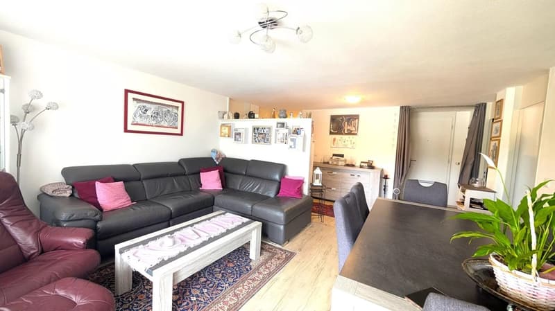 Spacious living room with a large L-shaped leather sofa, coffee table, and shelving unit with decorative items. The room has hardwood floors, white walls, and a modern light fixture. There is a balcony or terrace visible through the window.