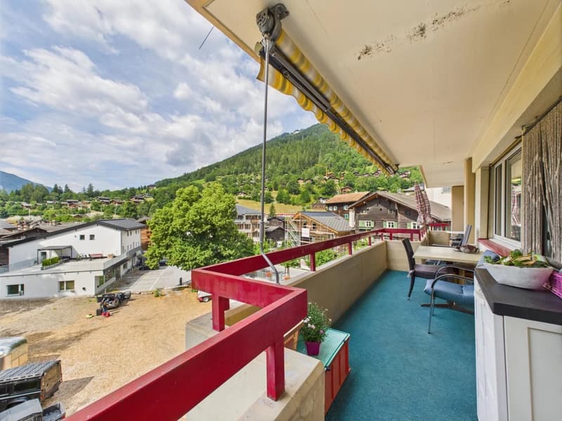 Balcony with a view of a mountainous, forested area and a town below. The balcony has a red railing and a retractable awning. The interior includes a seating area with chairs and a table.