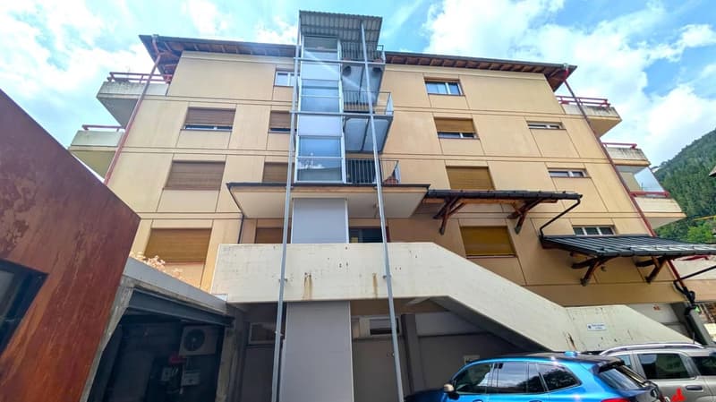 Multi-story apartment building with beige exterior, balconies, and a glass elevator shaft on the side.