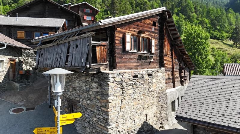 Traditional alpine-style wooden cabin with stone foundation, surrounded by lush green forest. The cabin has a balcony or terrace, and there are directional signs indicating nearby destinations.