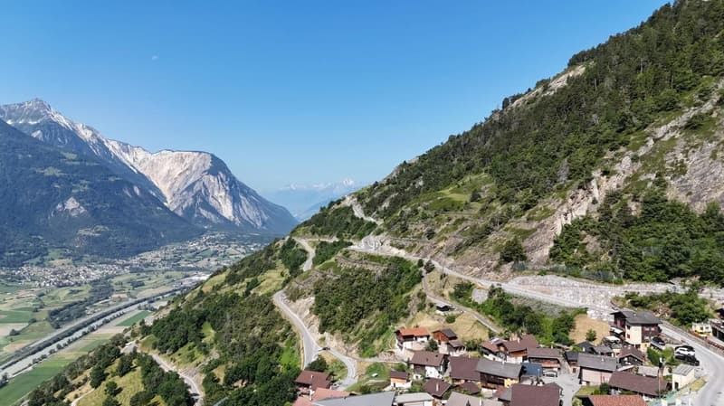The image shows a scenic mountain landscape with a winding road running through the valley. The mountains are covered in lush green forests, with rocky cliffs and peaks in the distance. A small village can be seen nestled in the valley below, with traditi