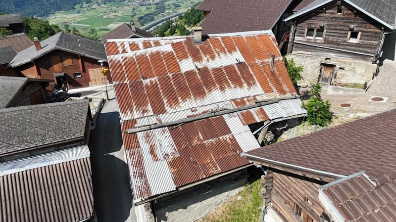 The image shows a rustic, old building with a corrugated metal roof. The roof appears to be in poor condition, with several damaged or missing sections. The building is surrounded by other similar structures, suggesting it is part of a small village or ru