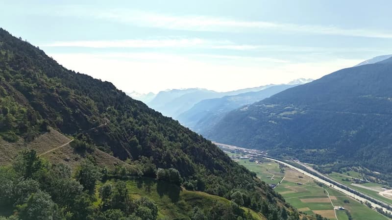 The image shows a scenic mountain landscape with lush green forests and valleys. The mountains rise up on both sides, creating a dramatic and picturesque view. There is a winding road visible in the foreground, leading through the valley below. The sky is