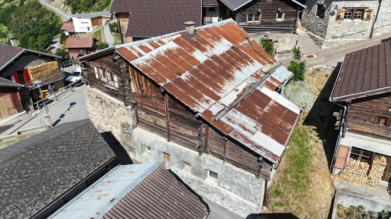 This image shows a traditional alpine-style wooden house with a stone foundation. The roof is made of rusty metal sheets, and the exterior walls are constructed with wooden logs. The house appears to be situated in a mountainous, forested area with other