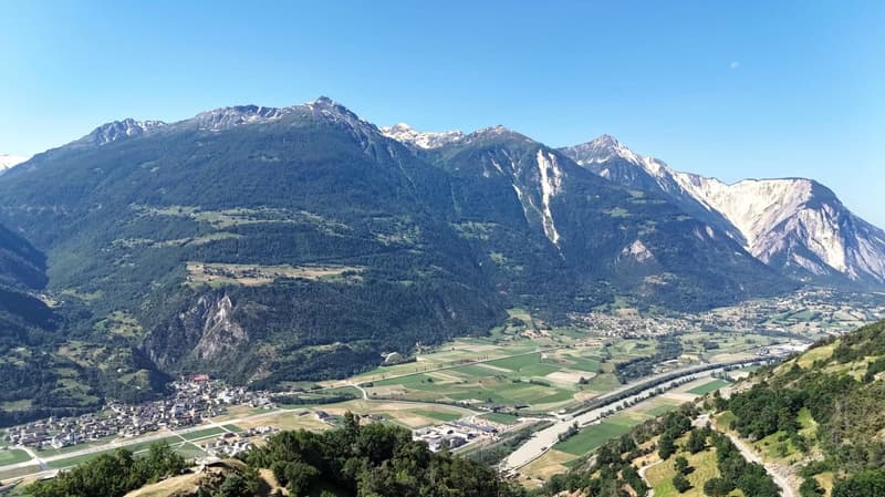 The image shows a stunning mountain landscape with snow-capped peaks, lush green valleys, and a small town nestled in the valley below. The scenery is breathtaking, with a clear blue sky providing a beautiful backdrop to the rugged, majestic mountains.