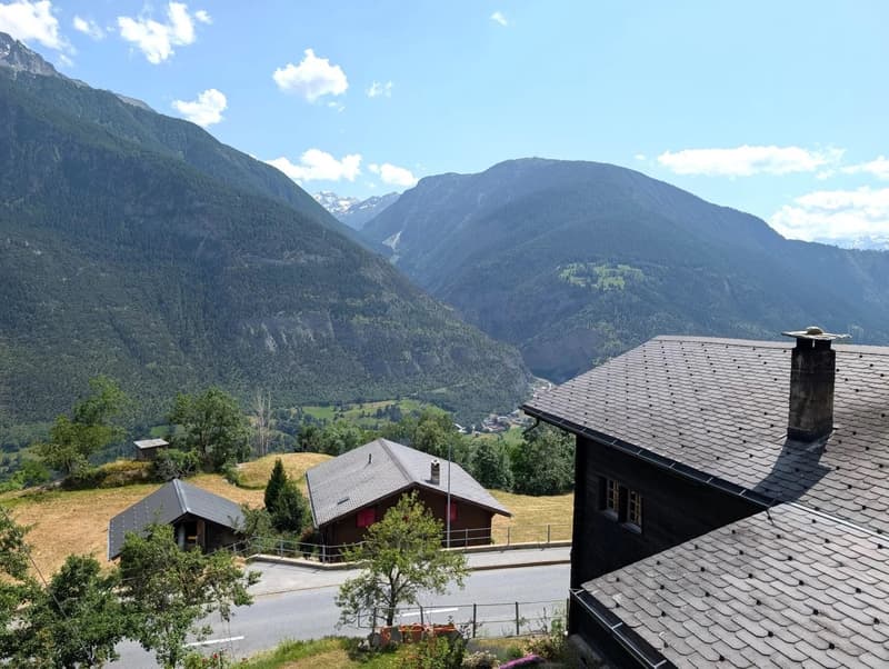 The image shows a scenic mountain landscape with a small village nestled in the valley below. The buildings have traditional alpine-style architecture with slanted roofs. There are several small cabins or chalets visible, surrounded by lush greenery and t