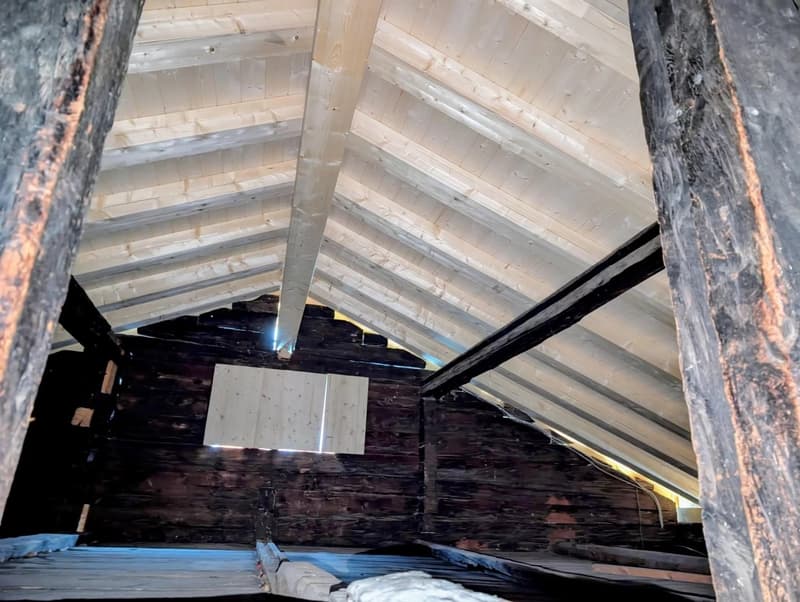 The image shows the interior of an attic space. The ceiling is made of wooden beams and boards, creating a rustic and exposed look. There are some wooden support structures visible, as well as what appears to be insulation or other building materials in t