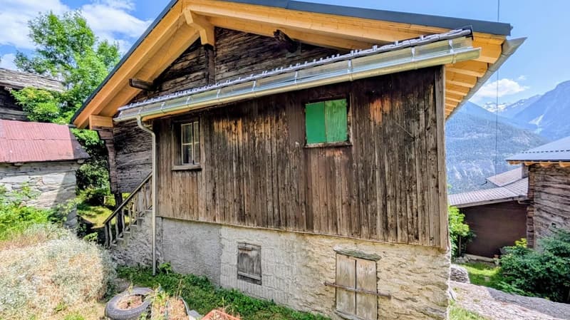 Rustic wooden cabin with a balcony, stone foundation, and a green window. The cabin is surrounded by lush greenery and mountains in the background.