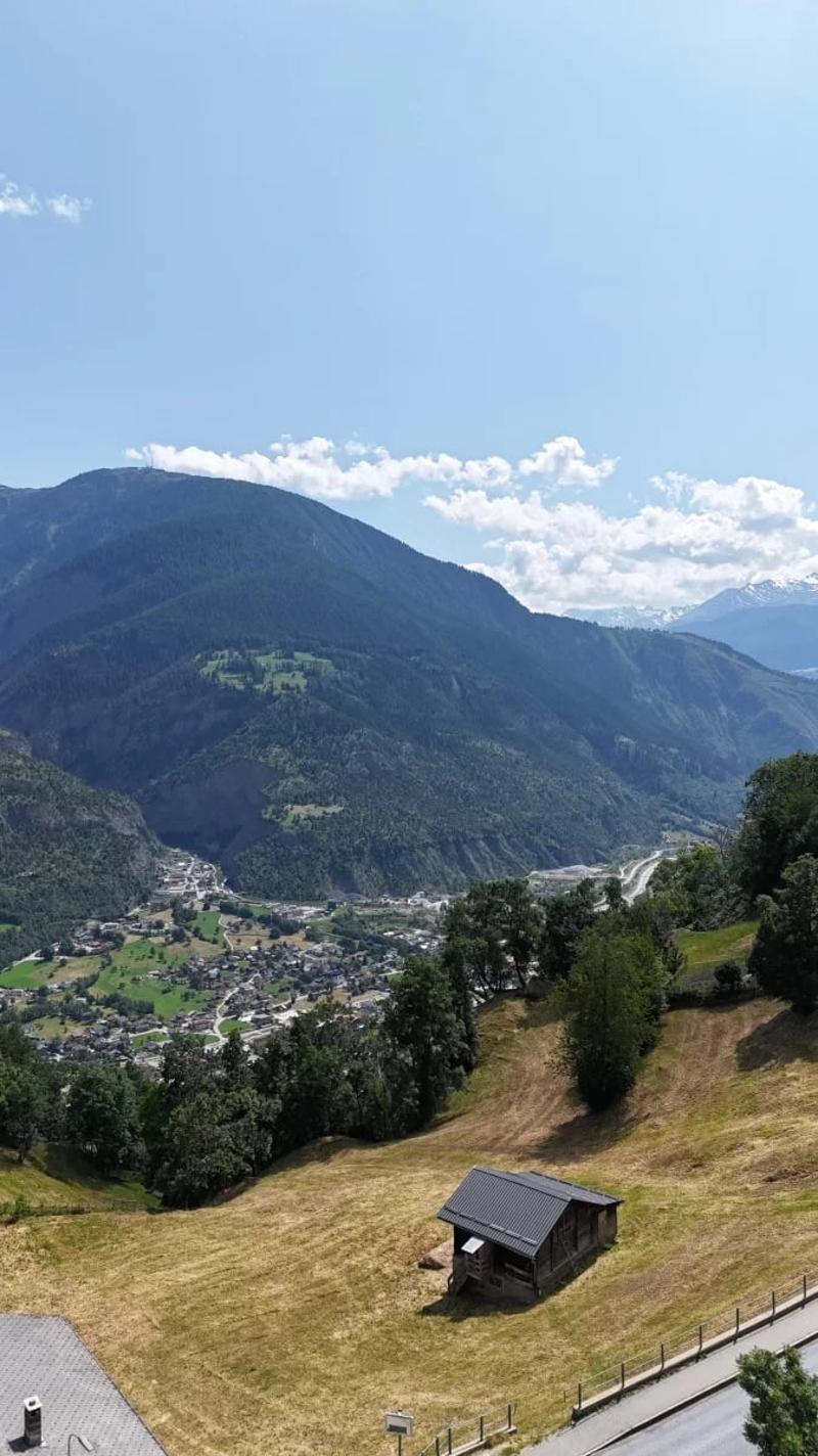 The image shows a scenic mountain landscape with a small village nestled in the valley below. The mountains are covered in lush green vegetation, with some snow-capped peaks visible in the distance. A small wooden cabin or shed can be seen on a grassy hil