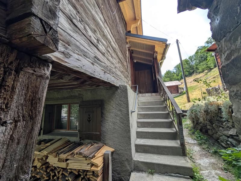 Rustic wooden cabin with a raised ground floor, stone steps leading up to the entrance, and a stack of firewood visible outside.