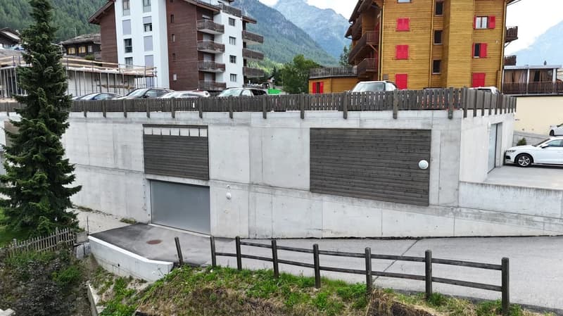 The image shows a multi-story residential building with a concrete and wooden exterior. There is a garage door visible on the ground level, indicating the presence of a garage. The building is surrounded by a wooden fence and is situated in a mountainous