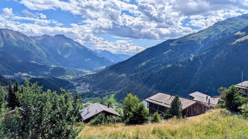 Two chalets, wooden exterior, several levels, surrounded by grassy slope, large mountains in the background, dense green forest, sunny weather with some clouds.
