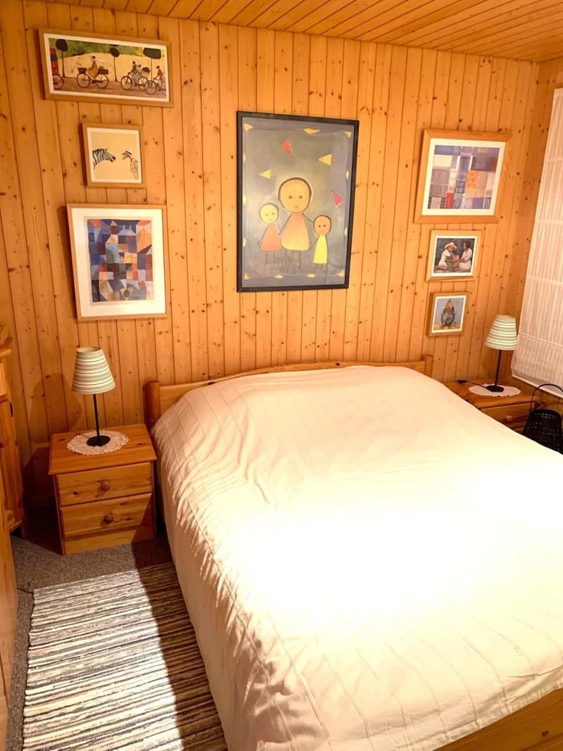 Wooden walls, bed with white bedding, two nightstands with lamps, a carpet, several framed pictures on the wall
