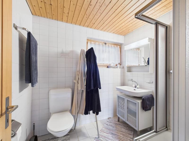 white tiles, white toilet, white sink, mirror, shower area, towel hanger, window with curtain, towel and robe on clothes rack