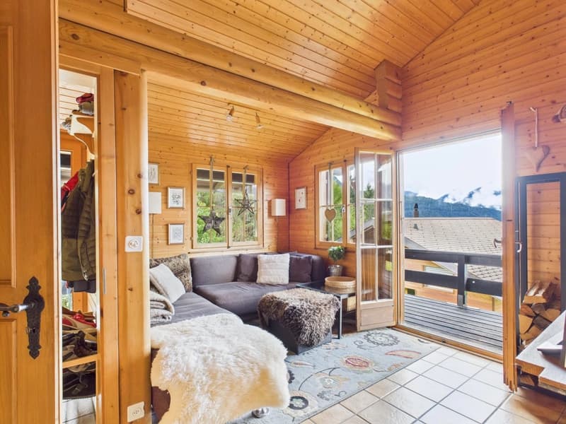 Log cabin styled interior, wooden walls, large couch, rug, sliding doors to balcony, glass window with view of mountains, fireplace