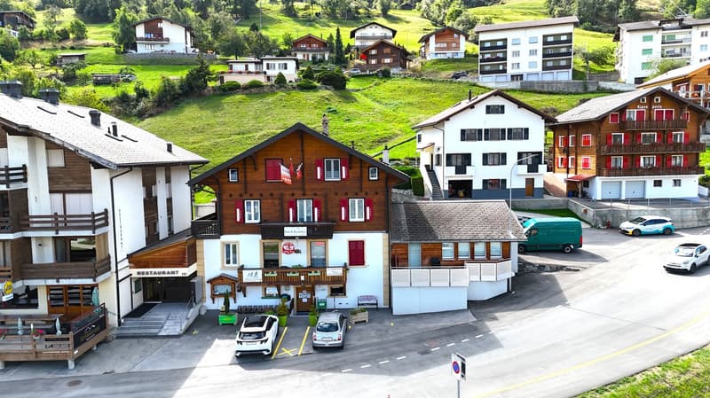 Mountain village complex with multiple buildings, wooden facades, and red shutters. One building has a restaurant sign. The complex is surrounded by a green hillside. Cars parked in front of the buildings.