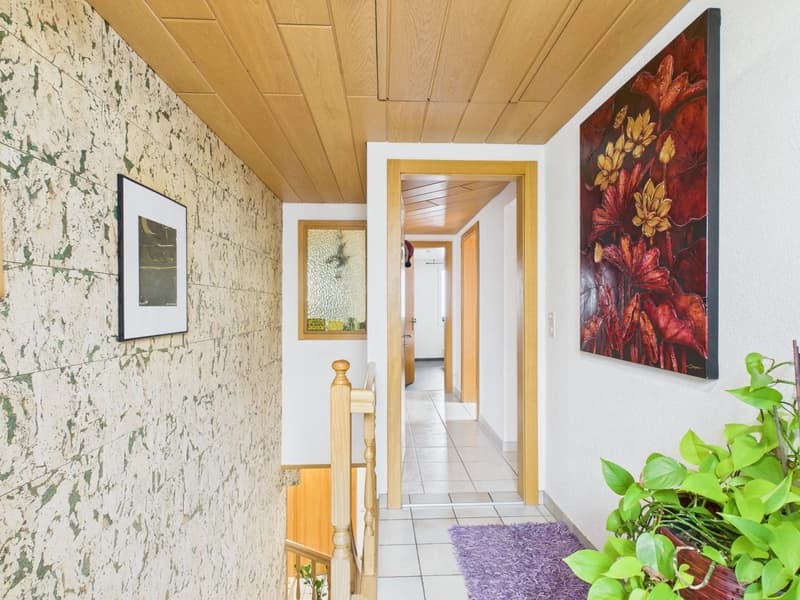 tiled floor, white walls, ceiling made of wood panels, wooden railing, painting on wall, photo frames, a rug, potted plant