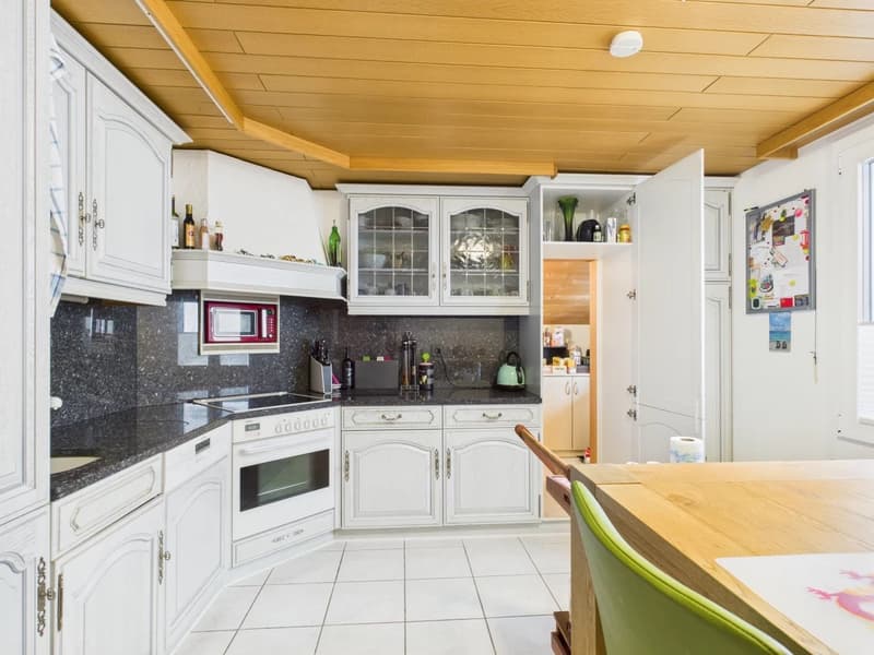 Modern kitchen with granite countertops, white cabinets, tiled flooring, wooden ceiling, gas stove, sink, microwave, coffee machine, teapot, various kitchen utensils, and a dining table.