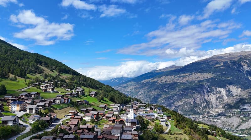 Town on a mountainside, surrounded by lush green meadows, mountains, a clear blue sky with scattered clouds