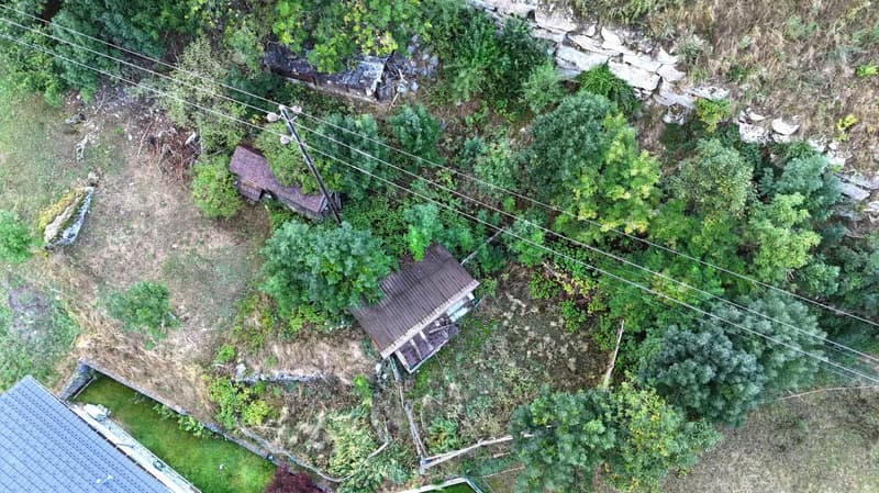 Overgrown land, several dilapidated houses, electrical poles and wires, large stone wall, surrounded by green vegetation