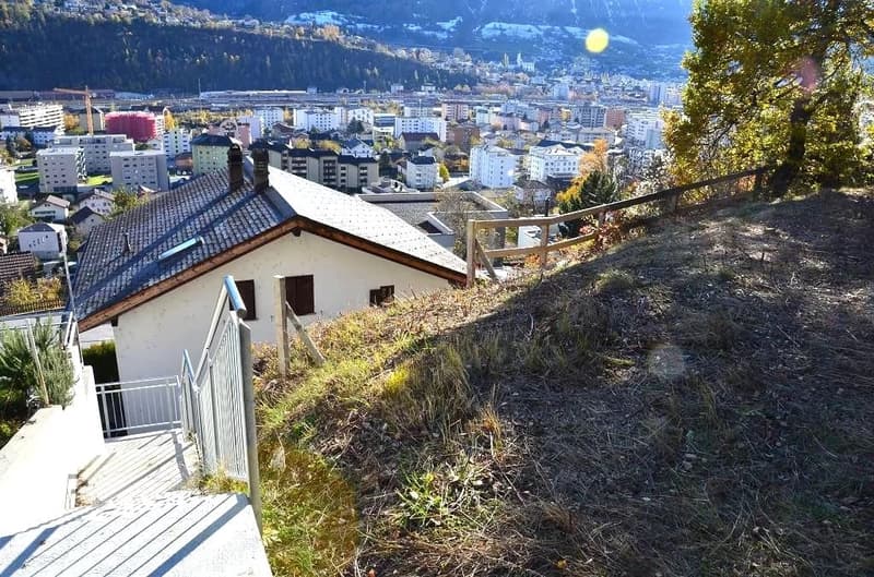 Slope-side house with steps leading up to it, surrounded by greenery and buildings in the distance, mountains in the background.