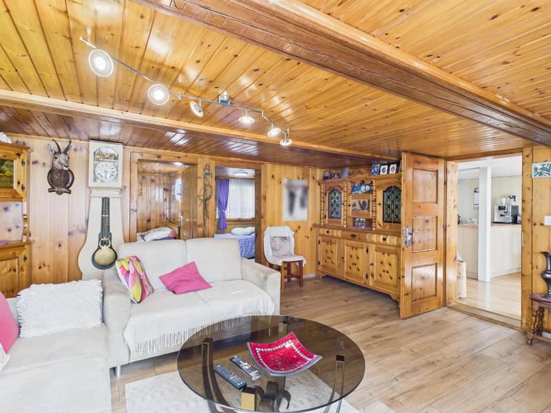 living room with wooden walls and floors, beige sofa with colorful pillows, round coffee table, wooden chair with pillow, vintage clock, and wooden cabinet with various items