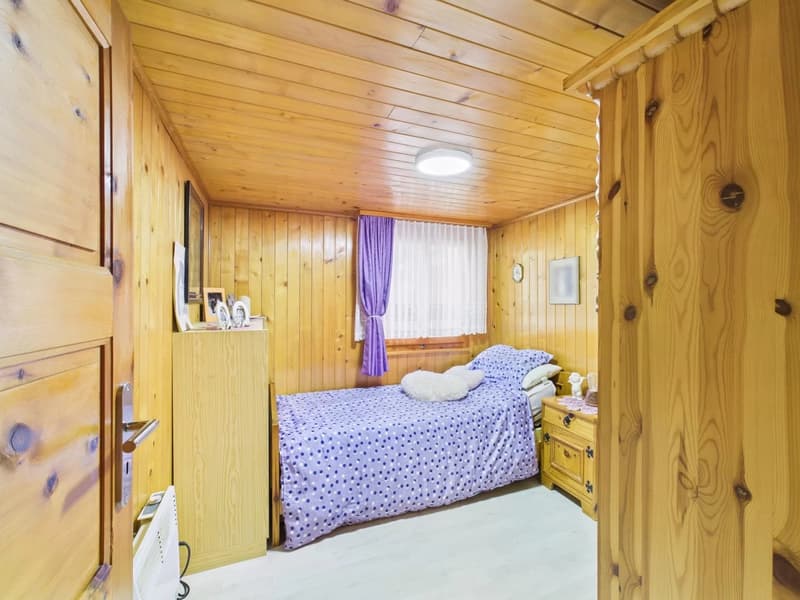 small bedroom with single bed, wooden walls, wooden floor, ceiling light, window with curtains, nightstand, bed, and wooden cabinet