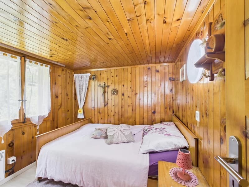 small bedroom, wooden walls, double bed, bed covers and pillows, nightstand, lamp, small cross on wall, small round window