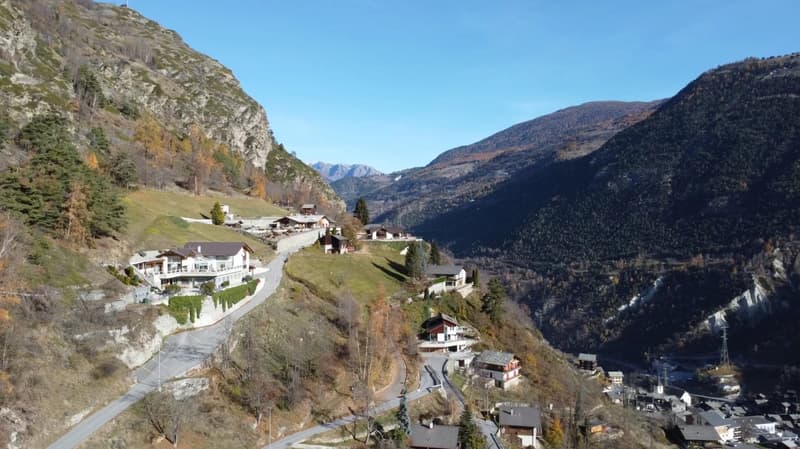 House exterior with a sloped road, mountainous area, trees, distant houses, mountains in the background