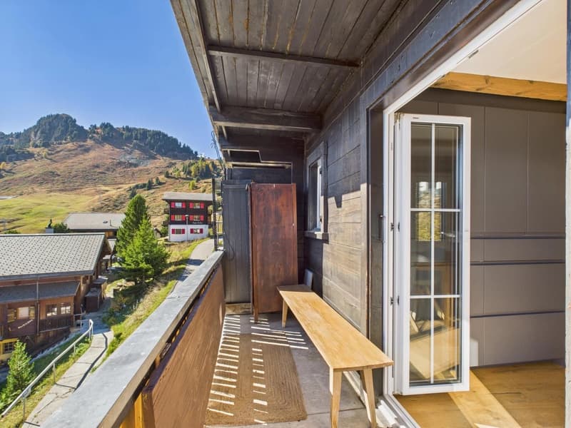 Exterior balcony of a building with wooden bench and door, located in a mountainous region