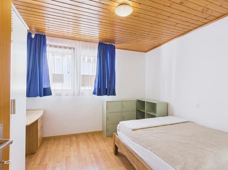 Small room with wooden floor, white walls, blue curtains, bed, wooden cabinet, light wooden ceiling