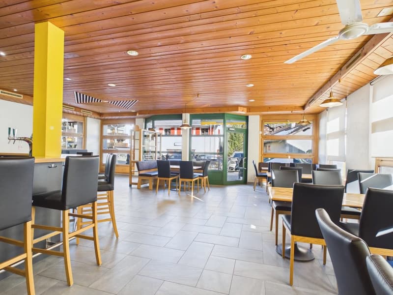 Interior of a restaurant with wooden benches, wooden tables, high ceiling with lamps, large windows, ceiling fan.