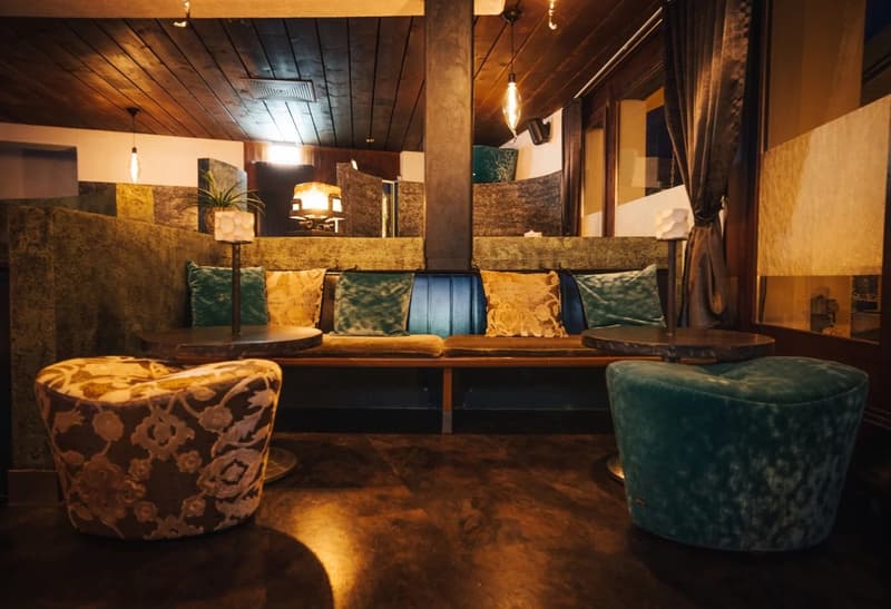 A cozy lounge area with a long bench, round tables, and two round chairs. The bench has multiple pillows, and the tables have potted plants on them. The room has wooden floors and a wooden ceiling with hanging lamps.