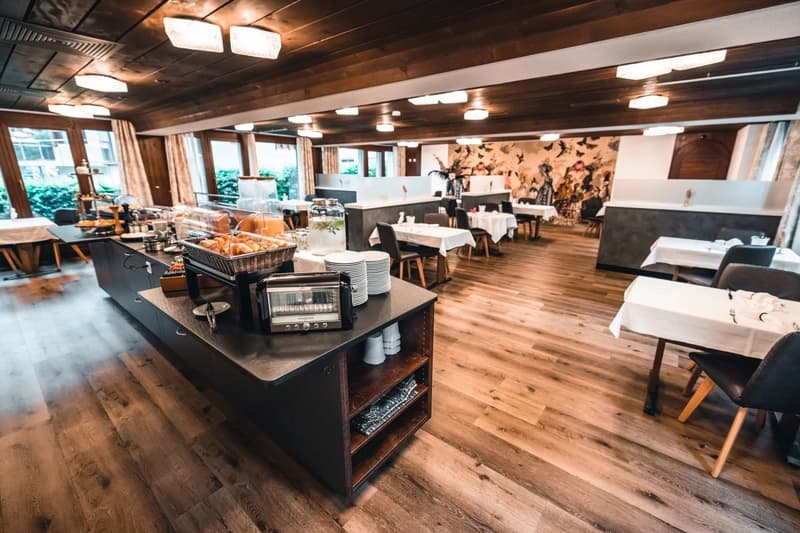 Wooden floor, wooden ceiling, large windows, buffet table, dining tables, chairs, plates, cups, toaster, bread, coffee machine, artwork on wall