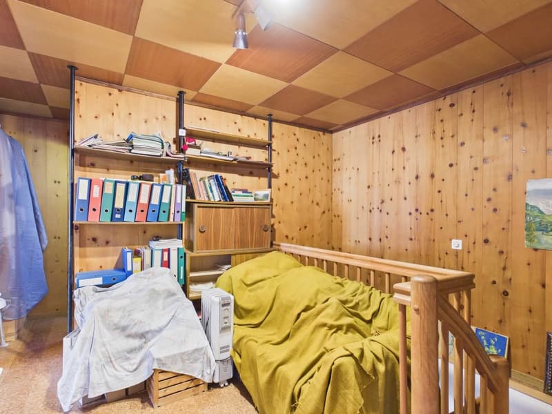 Small room with wooden walls, bed, wooden shelves, books, small heater, wooden bed frame, wooden staircase