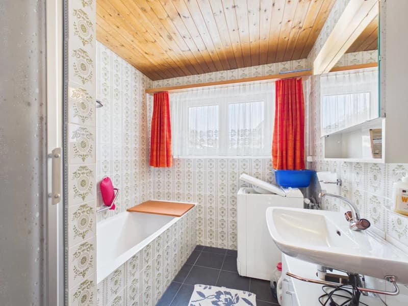 Bathroom with white bathtub, white sink, white washing machine, wooden ceiling, tiled walls, red curtains, and a mirror.
