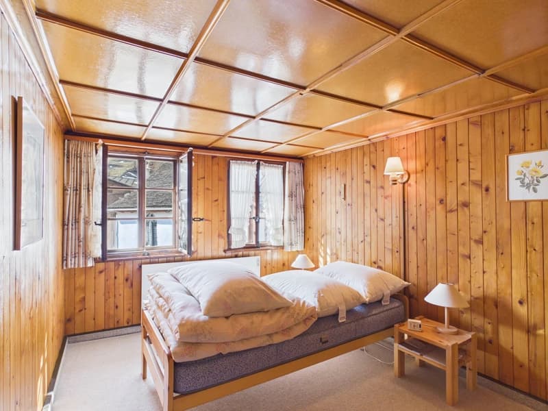 Bedroom with wooden walls, bed with pillows, two nightstands with lamps, windows with curtains, picture frames on the wall