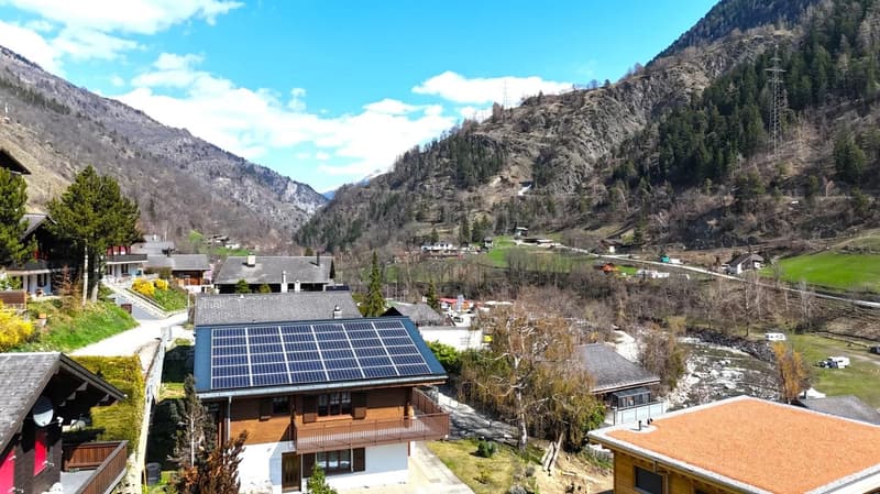House with solar panels on the roof, wooden balcony, surrounded by mountains and greenery