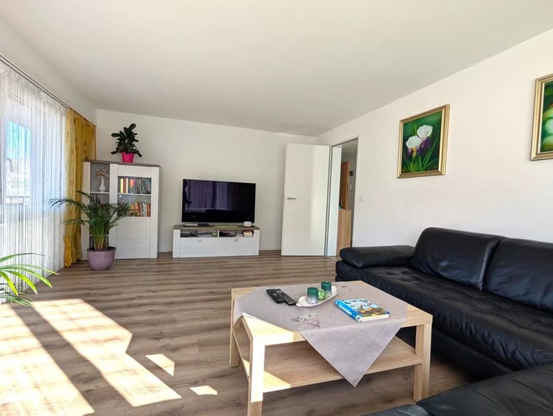 Living room with wooden floor, black leather couch, wooden coffee table, TV, potted plants, and framed pictures on the wall.