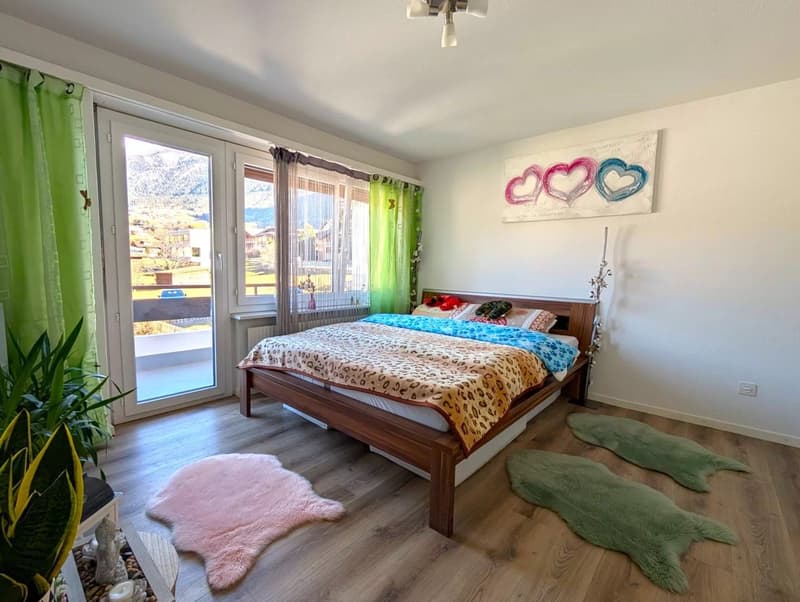 Bedroom with wooden bed, leopard print bedspread, two pink rugs, green rugs, green curtains, glass door leading to balcony, painting on wall, potted plant, wooden floor, ceiling lamp