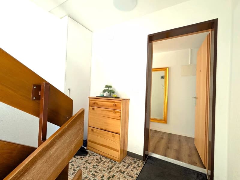 A hallway with wooden stairs, a wooden cabinet with drawers, a mirror, and a tiled floor.