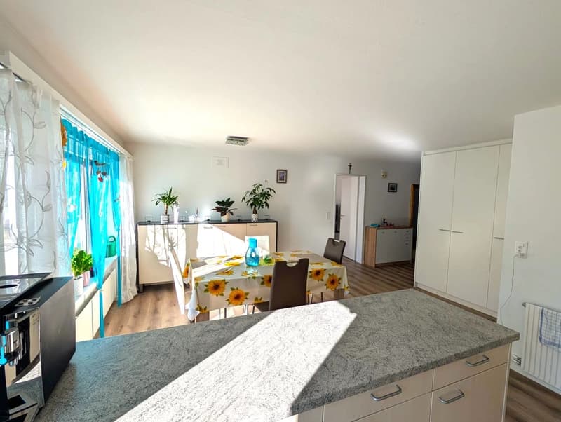 Dining room with a table and chairs, a counter with a coffee machine, a cabinet, and a window with blue curtains. Potted plants are on the window sill. The room has a wooden floor and white walls.