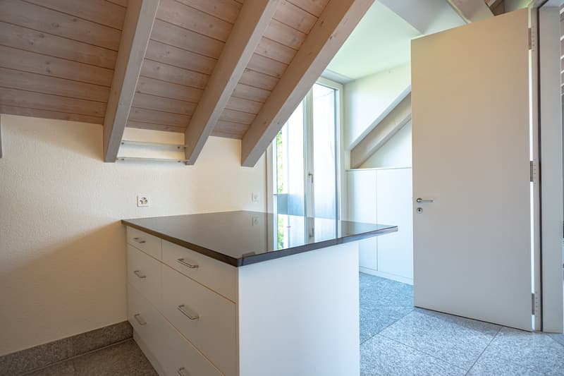 This image shows a room with a wooden ceiling, white walls, and a large window that provides natural light. There is a built-in cabinet or counter space with drawers, and the floor appears to be tiled or made of a stone-like material. The room has an open