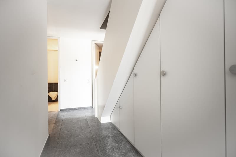 The image shows a hallway with white walls, a tiled floor, and built-in cabinets or closets along one side. The hallway leads to a doorway that appears to open into another room.
