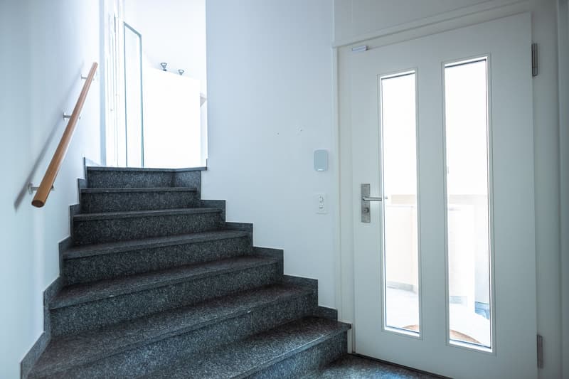 The image shows a staircase with gray granite steps leading up to a door. The walls are painted in a light color, and there are two small windows on the side providing natural light. The staircase appears to be the main entrance to the building.