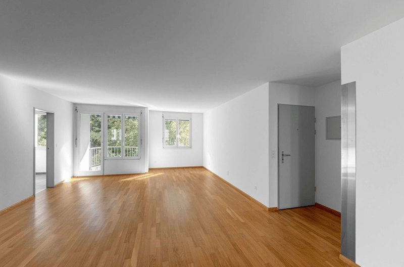 Large empty room with white walls, wooden floor, glass doors leading to balcony with railings