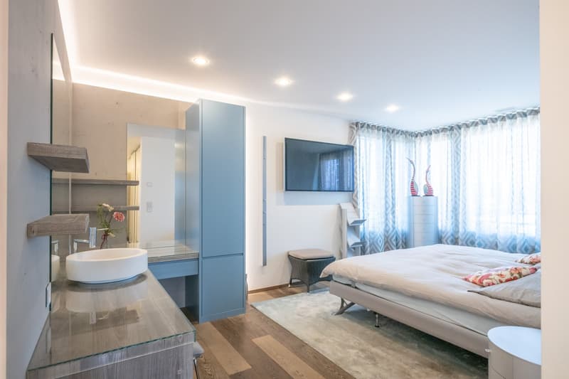 modern bathroom with oval sink, large mirror, wooden shelves, bedroom with bed, tv on wall, and white curtains
