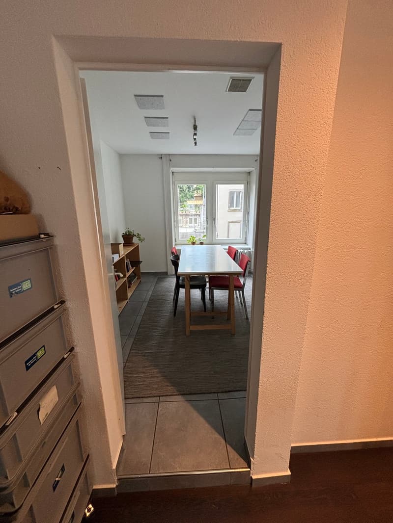 The image shows a dining room with a wooden table and chairs, shelves, and a window. The room has a tiled floor and white walls.