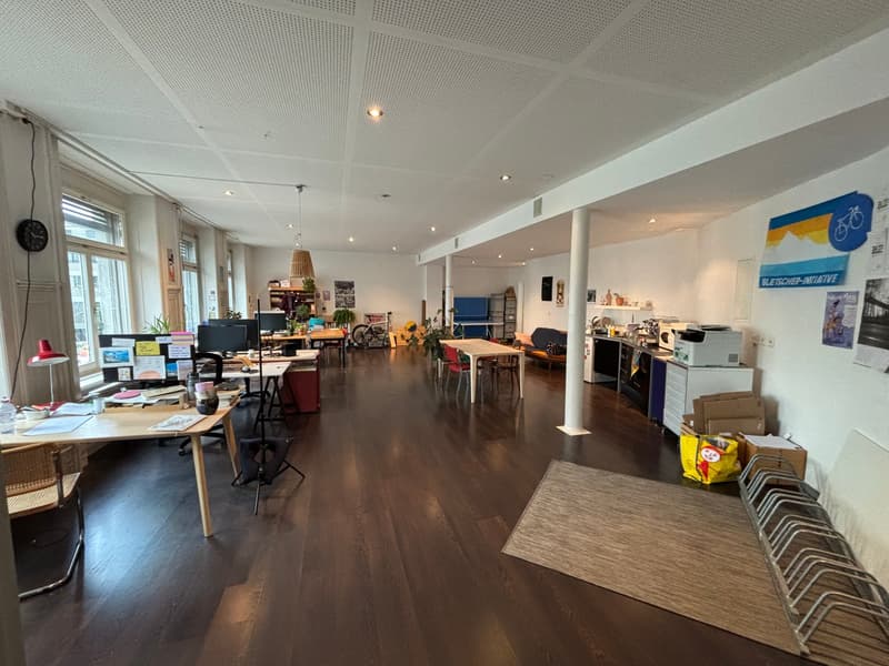 Large open-plan office space with hardwood floors, white walls, and a suspended ceiling. The space is furnished with desks, chairs, and various office equipment and supplies. There are also some personal items and decorations visible, suggesting it is a s