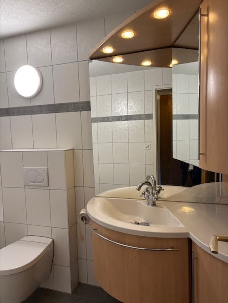 Bathroom with white tiles, a toilet, a sink, and a mirror with a wooden frame. There is a roll of toilet paper on the left side of the sink and a power outlet mounted on the wall above the toilet.